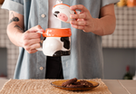 A cute ceramic mug shaped like a cow wearing an orange scarf and orange headphones. The cow has black and white spots and is sitting on a wooden surface. The mug has a whimsical and cozy design, perfect for fans of Lofi music and ChilledCow.