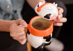 A cute ceramic mug shaped like a cow wearing an orange scarf and orange headphones. The cow has black and white spots and is sitting on a wooden surface. The mug has a whimsical and cozy design, perfect for fans of Lofi music and ChilledCow.