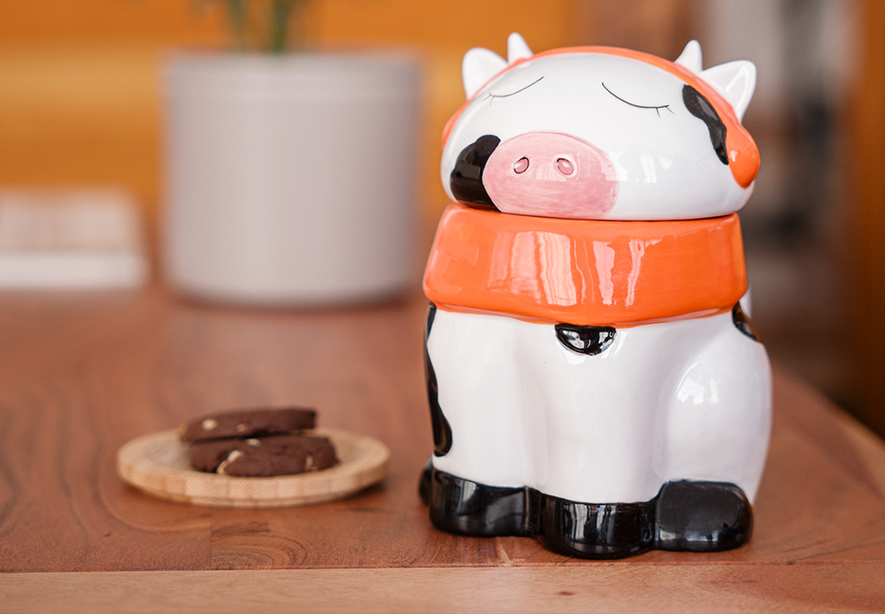 A cute ceramic mug shaped like a cow wearing an orange scarf and orange headphones. The cow has black and white spots and is sitting on a wooden surface. The mug has a whimsical and cozy design, perfect for fans of Lofi music and ChilledCow.