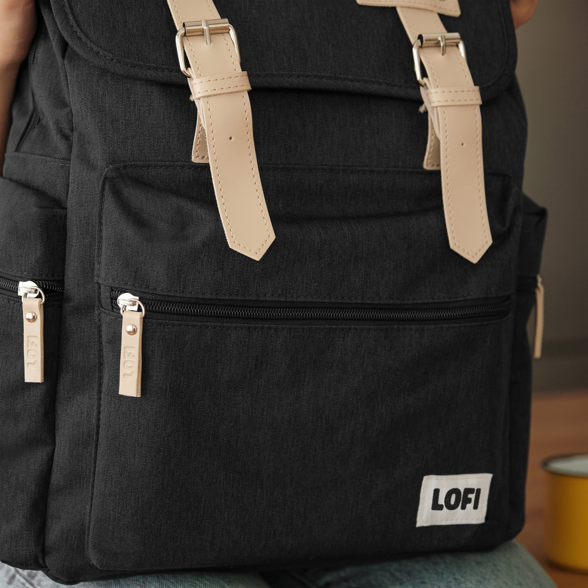 Close-up of a black Lofi Girl backpack, showcasing tan leather straps with metal buckles, and a front zipper pocket with leather pull tabs branded 'Lofi.' The backpack's minimalistic design, combined with its sturdy canvas material and stylish accents, emphasizes a functional yet fashionable aesthetic.