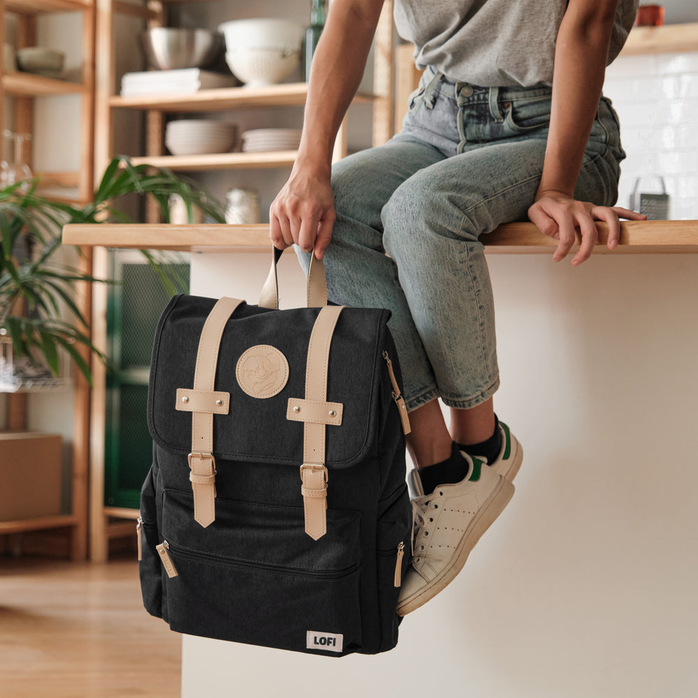 
                  
                    Person sitting on a kitchen counter, casually holding a black Lofi Girl backpack with tan leather straps and a circular embossed Lofi Girl patch. The room features open shelving with neatly arranged dishes and plants, providing a modern, homey backdrop to the stylish and functional backpack.
                  
                