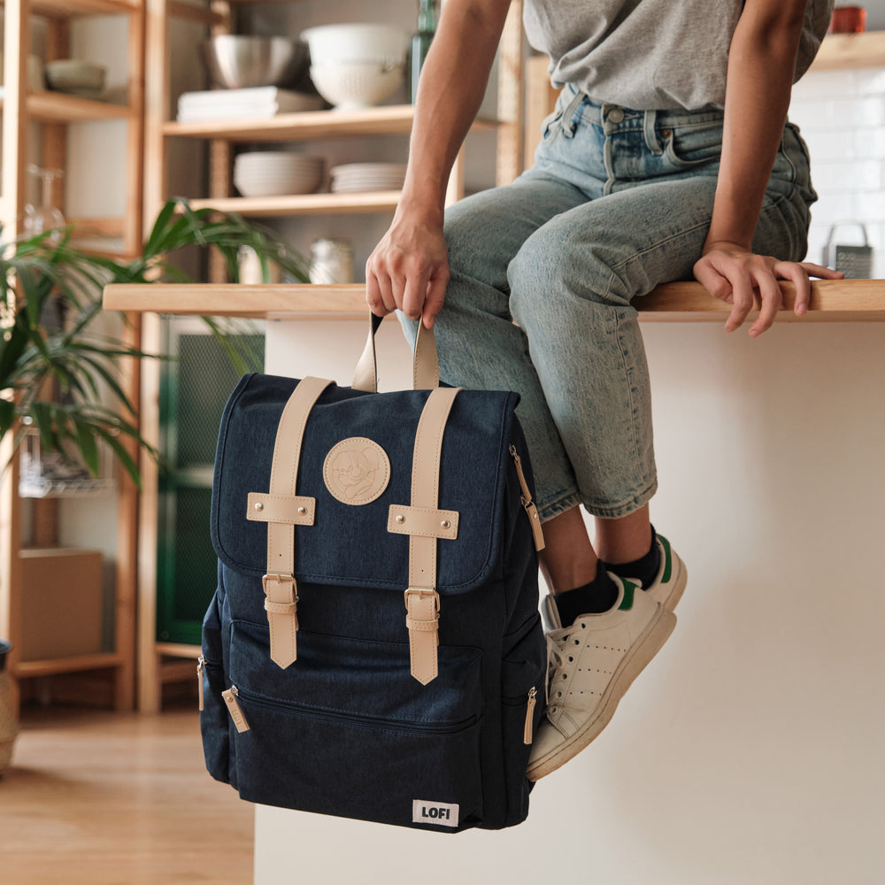 Person sitting on a kitchen counter, casually holding a navy blue Lofi Girl backpack with tan leather straps and a circular Lofi Girl patch. The room is styled with open shelves, potted plants, and a light, airy ambiance, creating a relaxed and contemporary vibe.