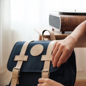 
                  
                    Close-up of a hand interacting with a navy blue Lofi Girl backpack, featuring tan leather straps and a circular embossed Lofi Girl logo on the flap. The background includes a vintage-style wooden turntable on a table, contributing to a warm and nostalgic atmosphere.
                  
                