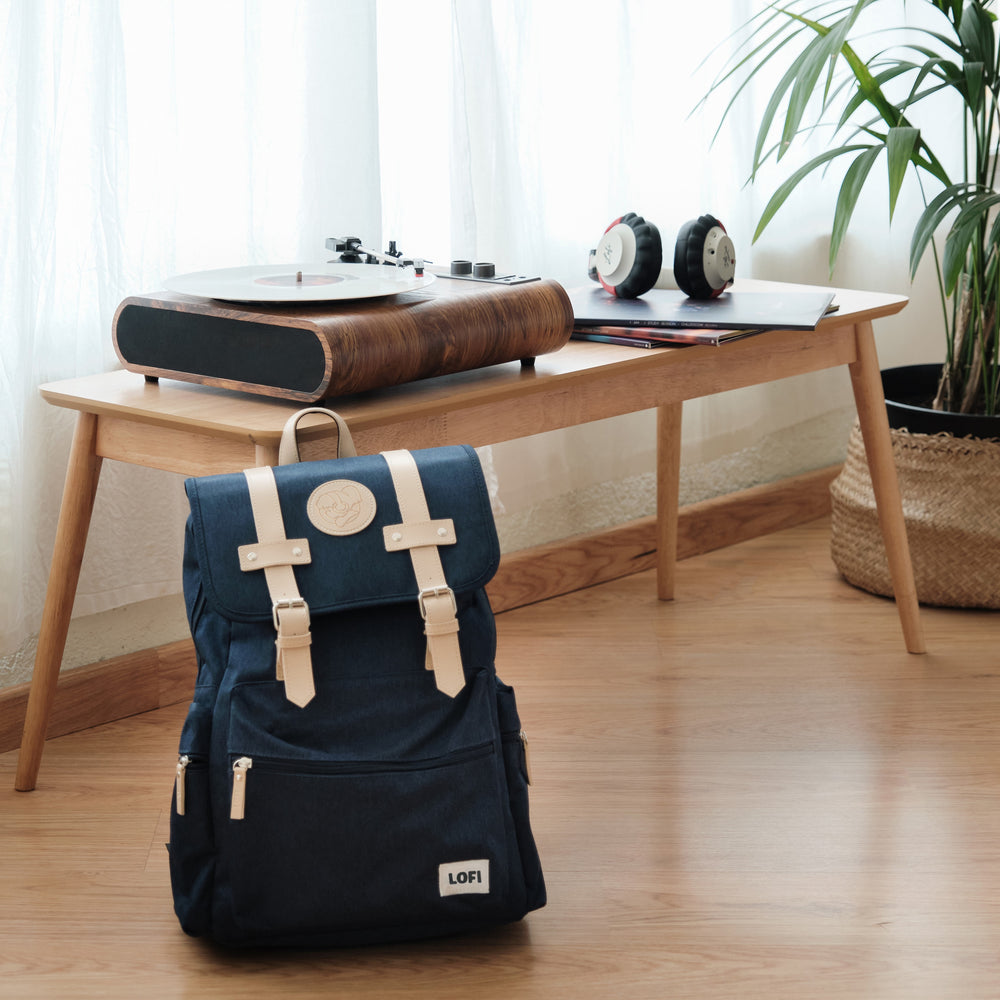 
                  
                    A stylish setup featuring a navy blue Lofi Girl backpack with tan leather straps and a circular logo, positioned on the floor in front of a wooden table. On the table is a vintage-style turntable, a pair of headphones, and some notebooks, with a potted plant adding a touch of greenery, creating a cozy and creative workspace ambiance.
                  
                