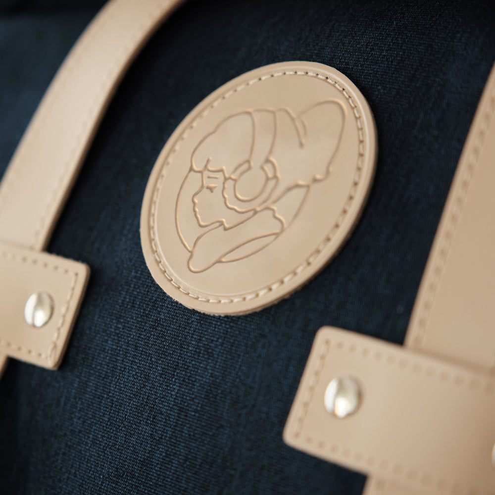 
                  
                    Close-up of the tan leather patch on a navy blue Lofi Girl backpack, featuring an embossed design of the Lofi Girl wearing headphones. The detailed stitching and rivet accents add to the quality craftsmanship and stylish design of the backpack.
                  
                