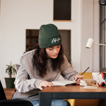 Person wearing a green beanie with the handwritten 'Lofi Girl' logo, paired with a grey sweatshirt, sitting at a wooden desk while focused on writing in a notebook. The setting is a cozy, well-lit room with minimalistic decor, including potted plants and a small desk lamp, creating a calm and studious atmosphere.