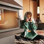 Cozy scene featuring a person sitting by a modern fireplace, wearing a green Lofi Girl plaid hoodie with a hood pulled over their head, smiling warmly. The room has a relaxed ambiance with soft lighting, wooden interior walls, and a fluffy rug on the floor.