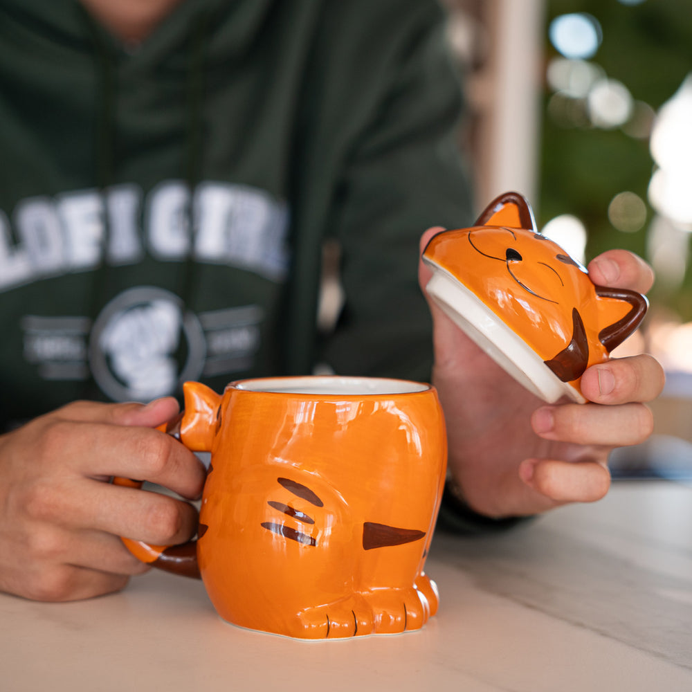 
                  
                    A person wearing a green hoodie with the "Lofi Girl" logo is holding an orange ceramic cat mug with a removable lid shaped like a cat's smiling face. The mug is designed to resemble a cute, cartoonish cat with stripes on its body.
                  
                