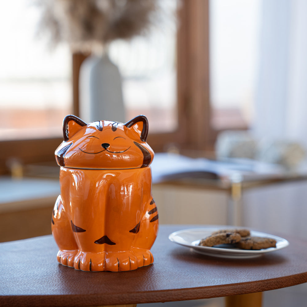 
                  
                    An orange ceramic cat mug with a smiling cat face lid sits on a wooden table. The background shows a cozy indoor setting with a blurred vase and some cookies on a plate nearby.
                  
                