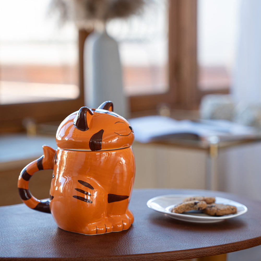 
                  
                    Close-up of an orange ceramic cat mug with a cat face lid on a wooden table. The mug has a tail-shaped handle, and a plate of cookies is placed beside it. The background features a cozy room with soft natural light.
                  
                