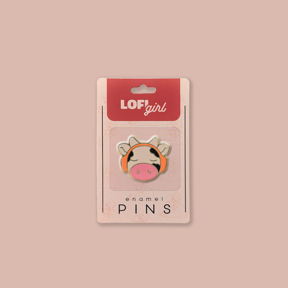 Lofi Girl enamel pin featuring a cute cow character wearing orange headphones, eyes closed in a relaxed expression. Packaged on a pink backing card with 'Lofi Girl' branding.