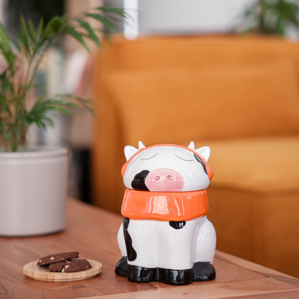 A cute ceramic mug shaped like a cow wearing an orange scarf and orange headphones. The cow has black and white spots and is sitting on a wooden surface. The mug has a whimsical and cozy design, perfect for fans of Lofi music and ChilledCow.