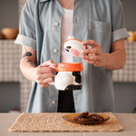 A cute ceramic mug shaped like a cow wearing an orange scarf and orange headphones. The cow has black and white spots and is sitting on a wooden surface. The mug has a whimsical and cozy design, perfect for fans of Lofi music and ChilledCow.