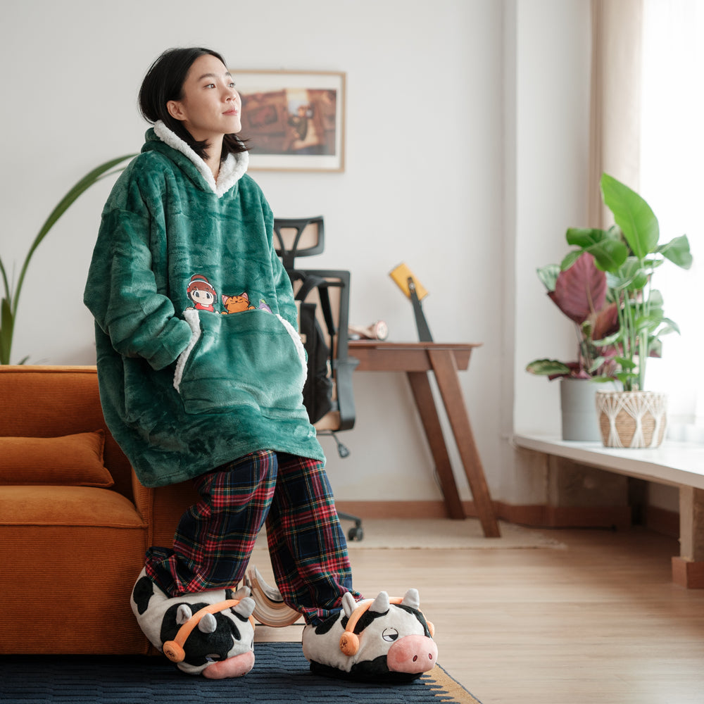 A woman is sitting on an orange sofa, wearing a green plush hoodie with an embroidered design of a cartoon character and cat. She has on plaid pajama pants and novelty slippers shaped like cows wearing headphones. The scene conveys a cozy and relaxed atmosphere in a well-lit living room.