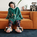 The same woman stands in a living room, wearing a green plush hoodie with cartoon embroidery and plaid pajama pants. She is also wearing cow-shaped novelty slippers with headphones. She appears to be looking out the window, with the room's interior, including plants and a desk, visible in the background. The setting suggests a comfortable, casual environment.