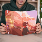 A close-up view of the Daytime Bullet Journal being held by a person wearing a green hoodie. The cover art features a serene scene of a Lofi Girl listening to music on a balcony under a bright, sunlit sky with puffy clouds. The illustration showcases a charming mix of urban and natural elements, with potted plants and a contented atmosphere.