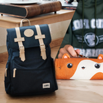 A navy blue backpack with beige straps and a Lofi Girl logo is positioned on the floor near a modern desk. Beside it, a hand holds an orange pencil case shaped like a cute animal, adding a playful touch to the setup.