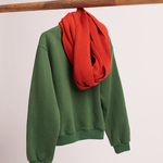 A green sweatshirt hangs from a wooden bar with a vibrant red scarf looped around the neck area. The sweatshirt and scarf are part of the iconic Lofi Girl's outfit.