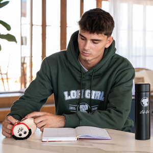 
                  
                    A young man is depicted in a relaxed study setting, interacting with the headphone pencil case. He appears engaged and thoughtful, flipping through a notebook while the pencil case sits nearby, adding a touch of style to the scene. The student is wearing a dark green hoodie with a "Lofi Girl" logo, further tying into the theme. A black thermos with the same branding stands on the table, suggesting a warm and focused study atmosphere.
                  
                