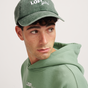 
                  
                    A close-up of a person wearing a vintage green cap with the "Lofi girl" logo embroidered in white on the front. The person has a focused expression and is looking to the side, dressed in a matching green hoodie. The cap has a slightly faded, vintage style that complements the casual, relaxed look.
                  
                