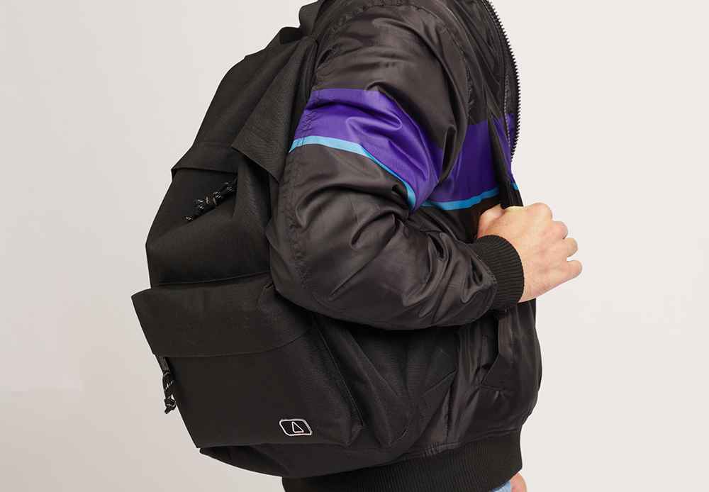 A person wearing a black jacket with purple and blue stripes carries a black "Lofi Boy" backpack on one shoulder, with their right hand holding the shoulder strap. The backpack features a simple, minimalist design with a small logo on the front pocket. The background is plain and neutral.