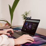 A side view of a desk setup showcasing a person using a laptop on a "Lofi Boy" desk pad. The animated desk pad shows a character typing at a computer in a serene nighttime scene. In the background, there are plants and other Lofi-themed merchandise.