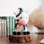 A charming 'Medieval ChilledCow' plush sits on a rustic wooden platform, dressed in a colorful jester-like outfit with bells on its feet and a hat adorned with a gold bell. The plush features whimsical star patterns on its face, adding a playful touch. In the background, there is a collection of classic books and a vintage globe, creating a cozy and nostalgic ambiance that perfectly complements the medieval theme of this unique collectible.