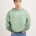 A young man with curly black hair wearing a sage green "Lofi Girl" hoodie with his hands in the front pocket, standing against a plain background.