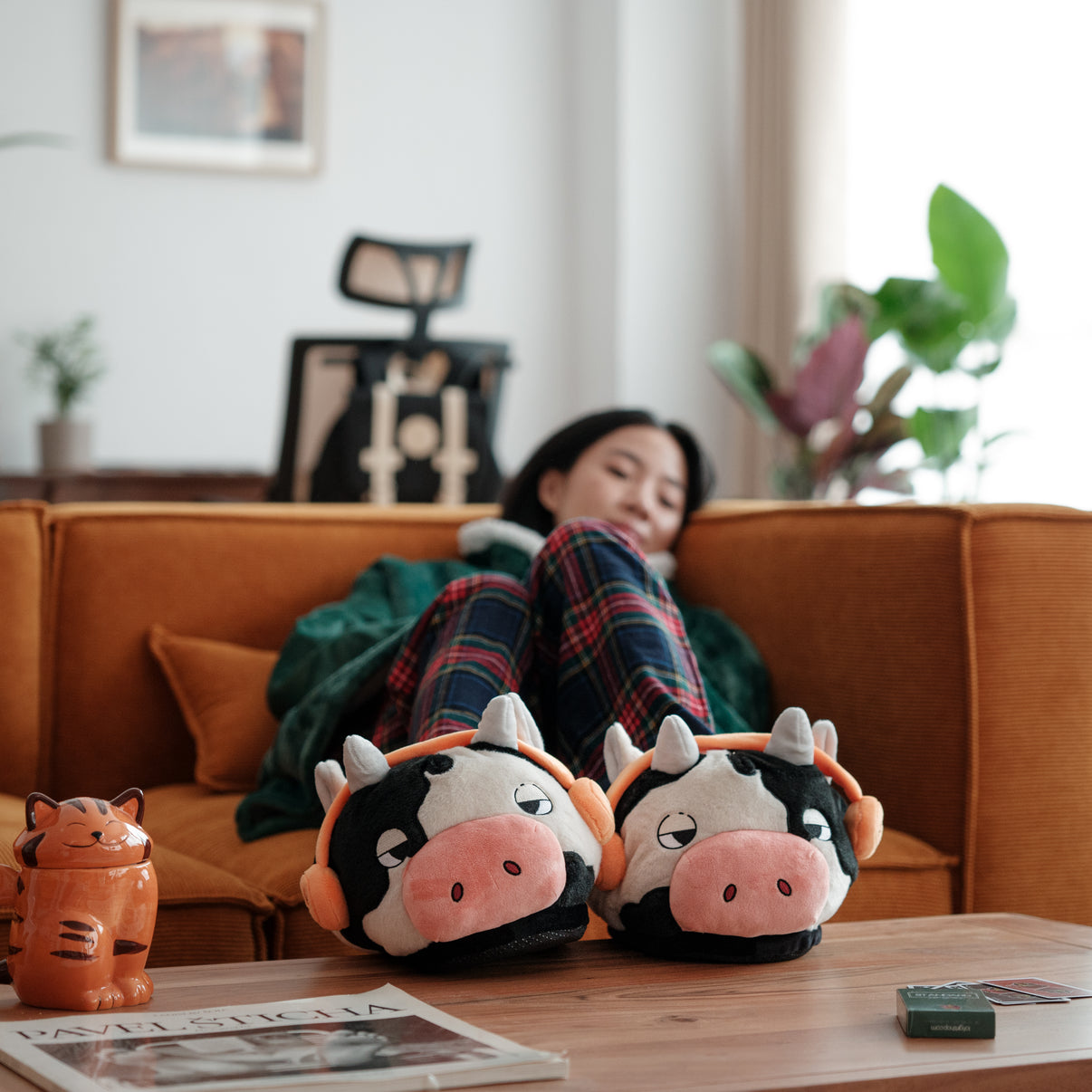 A woman lounging on an orange sofa wearing Lofi Girl slippers shaped like cows with headphones. The slippers are prominently displayed on a coffee table, with a cozy and relaxed living room setting in the background.
