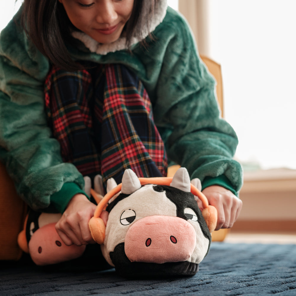 
                  
                    A close-up of a woman’s hands adjusting the Lofi Girl slippers shaped like cows with headphones, emphasizing the comfort and whimsical design of the slippers.
                  
                