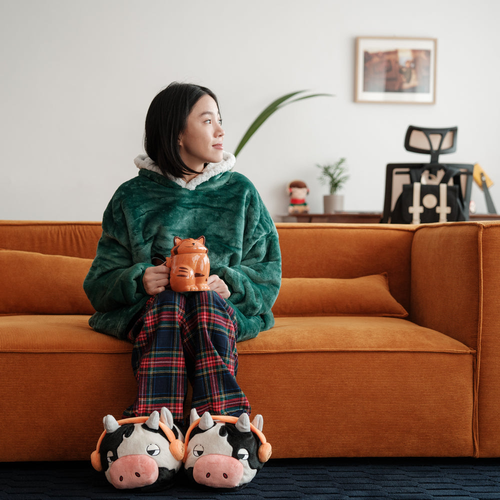 
                  
                    A woman sitting on an orange sofa, holding a mug shaped like a cat, wearing Lofi Girl slippers shaped like cows with headphones. She looks thoughtful, surrounded by a cozy and warm living room setting.
                  
                