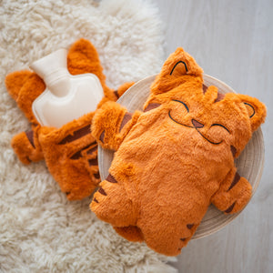 
                  
                    A close-up of an orange plush hot water bottle cover designed to look like a sleeping cat. It is placed on a wooden stool, with a soft, furry rug in the background.
                  
                