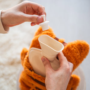 
                  
                    Hands are seen removing the stopper from a hot water bottle, which is partially inserted into the orange plush cover designed like a sleeping cat. The scene is set against a soft, light-colored rug, creating a cozy atmosphere.
                  
                