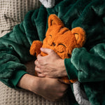 A person dressed in a dark green fleece robe is hugging the orange plush hot water bottle cover designed to look like a sleeping cat. The setting is cozy, with the person appearing relaxed and comfortable, emphasizing the comforting nature of the hot water bottle cover.