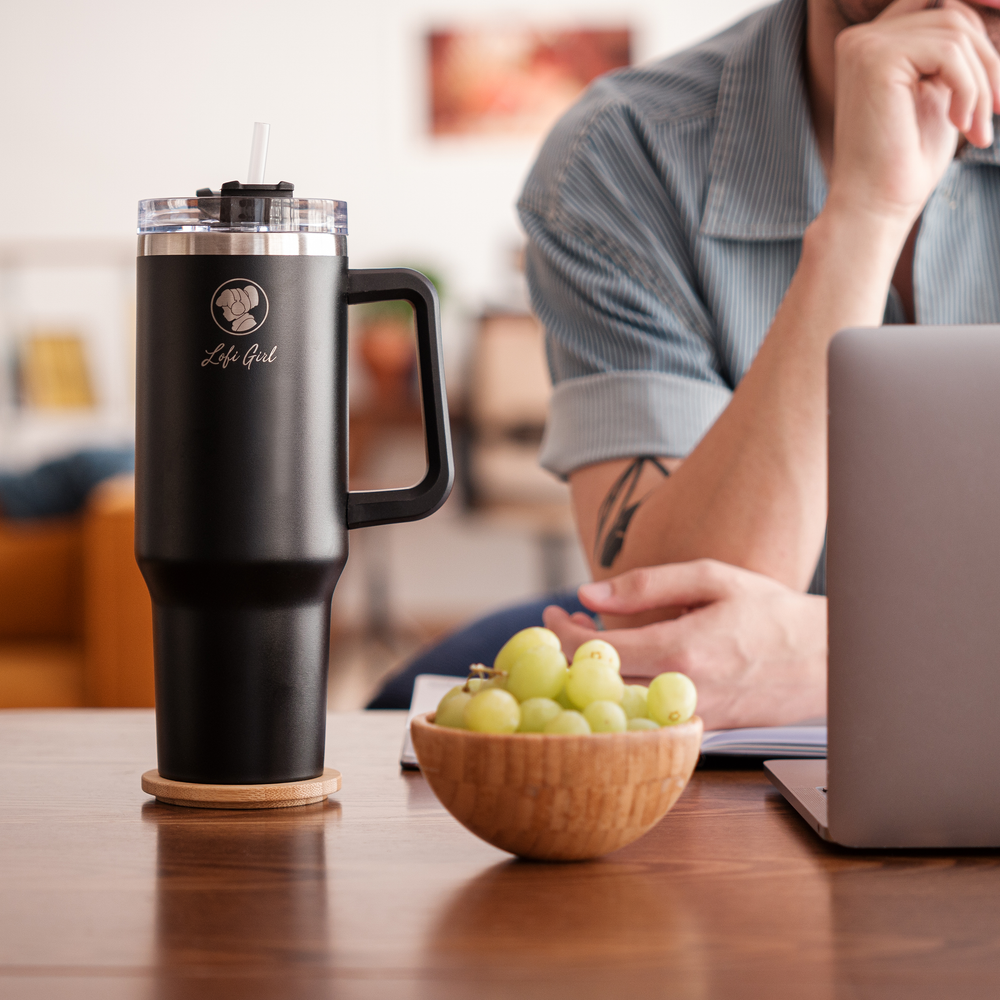 
                  
                    The Lofi Girl tumbler is placed on a wooden table next to a bowl of green grapes. A person in the background is sitting at the table with a laptop, with their hand near their face in a thoughtful pose.
                  
                