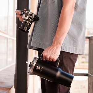 
                  
                    A person holding a camera in one hand and the Lofi Girl tumbler in the other. The scene is set in an outdoor area with sunlight filtering through, highlighting the tumbler's design and practicality for on-the-go use.
                  
                