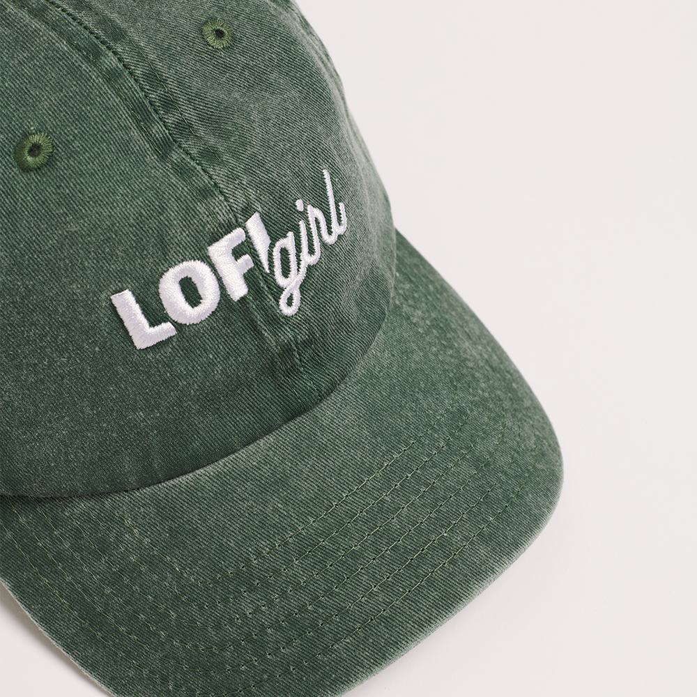 
                  
                    A close-up angled view of the front of a vintage green cap. The focus is on the embroidered "Lofi girl" logo in white on the front panel, with the curved brim visible at the bottom.
                  
                