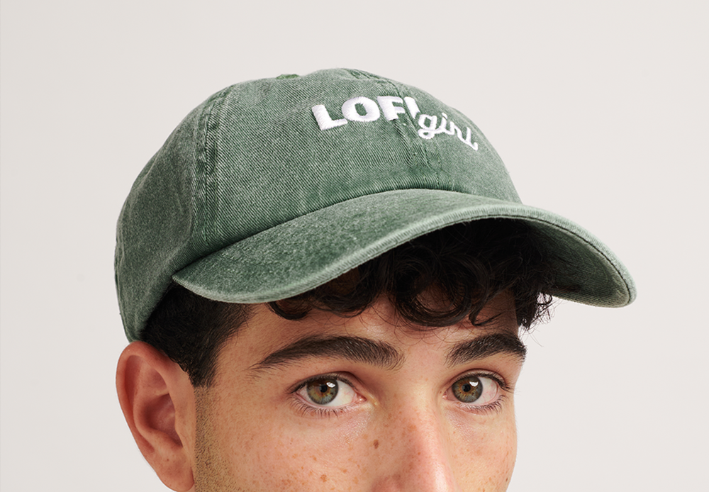 A portrait of a person wearing a vintage green cap with the "Lofi girl" logo embroidered in white on the front. The person is looking slightly to the side, and the cap has a faded, vintage style that complements the casual outfit.