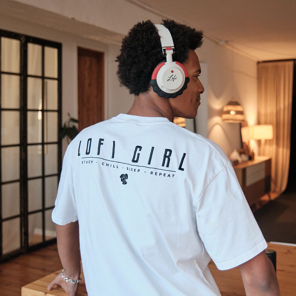 Person wearing an oversized white Lofi Girl t-shirt with a bold black 'Lofi Girl' logo on the back, paired with the tagline 'Study • Chill • Sleep • Repeat.' They are wearing headphones, standing in a warmly lit, stylish interior with a relaxed and cozy vibe.