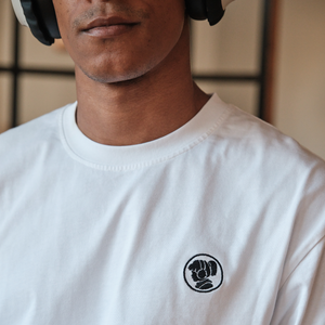 
                  
                    Close-up view of a person wearing a white Lofi Girl t-shirt, featuring a small, circular embroidered logo on the chest depicting a stylized fist. The person is also wearing headphones, partially visible in the image, creating a minimalist and modern look.
                  
                