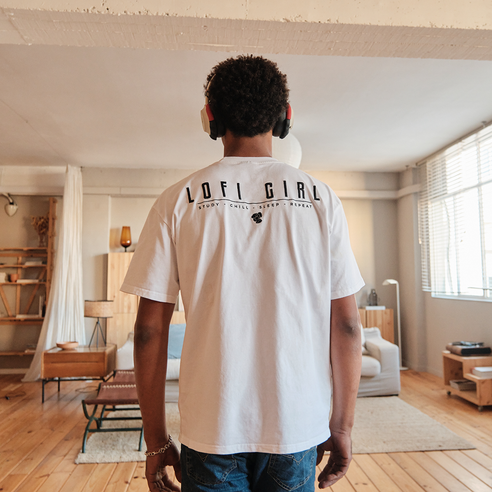 
                  
                    Person standing in a spacious, modern living room, wearing a white Lofi Girl t-shirt with bold black lettering on the back that reads 'Lofi Girl' along with the tagline 'Study • Chill • Sleep • Repeat' and a small fist icon. They are wearing headphones, adding to the relaxed and comfortable atmosphere of the room.
                  
                