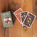 A deck of Lofi Girl playing cards is spread out on a wooden table. The card box features an illustration of the Lofi Girl character with her cat and a frog. Three cards are partially visible: a red King of Diamonds with a cat illustration, a black King of Clubs with Lofi Girl, and a black King of Spades with a frog character.