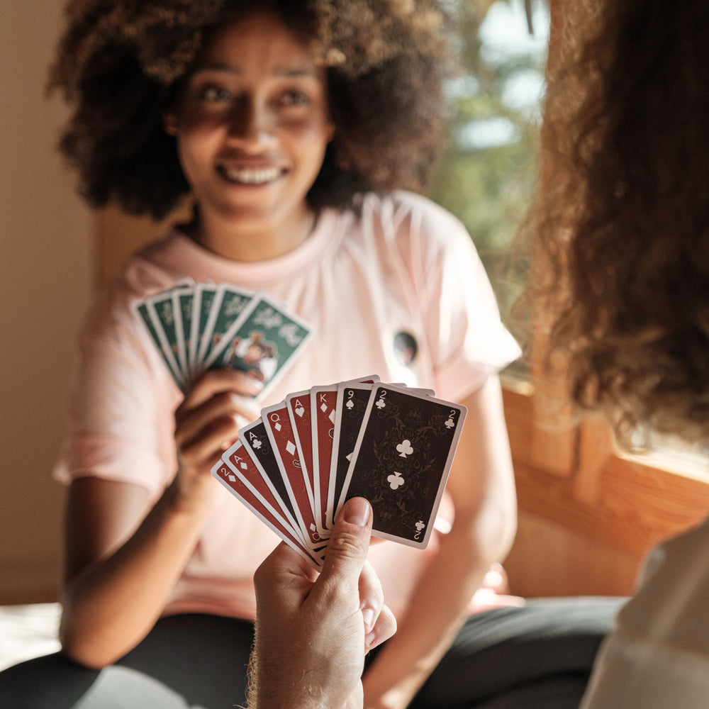 
                  
                    Two people are playing with Lofi Girl-themed playing cards. A person in the foreground holds a fan of cards, including a King of Clubs and a King of Spades with illustrations of Lofi Girl characters. The other person, wearing a pink shirt, smiles while holding their cards in a spread.
                  
                