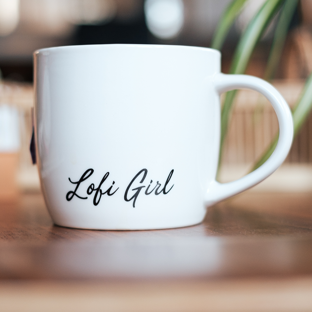 
                  
                    Close-up of a white ceramic mug displaying the cursive text "Lofi Girl" on one side. The mug is positioned on a wooden table, and a plant is slightly visible in the background, creating a cozy, warm ambiance.
                  
                