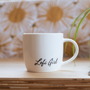 
                  
                    A white ceramic mug with the words "Lofi Girl" in cursive on the side. The mug is placed on a light-colored wooden surface, with a blurred floral-patterned background and a green plant leaf visible on the right.
                  
                